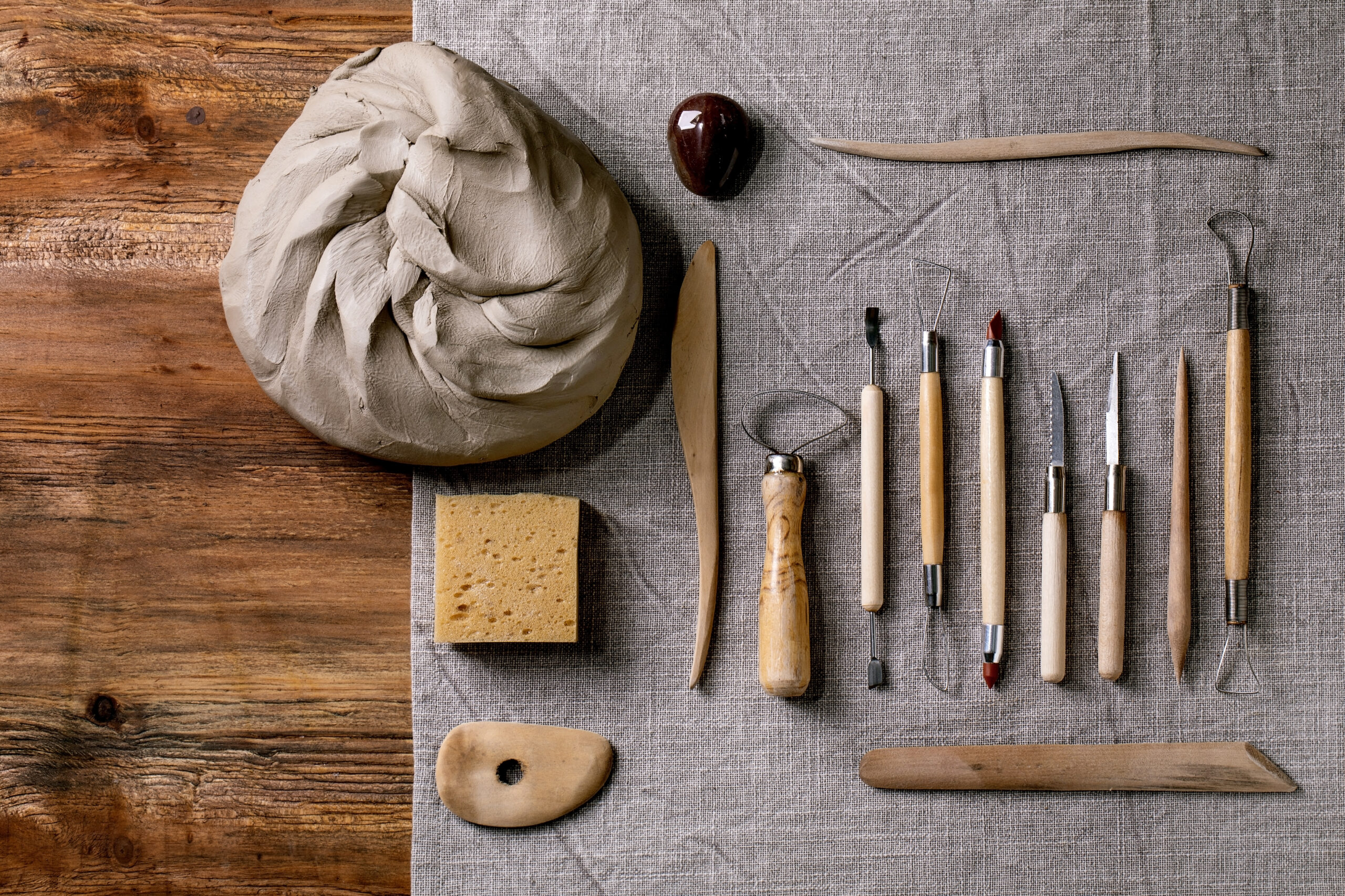 Set for pottery hobby. Piece of raw grey clay and kit of pottery ceramics tools in row on linen fabric cloth. Top view. DIY potter hobby
